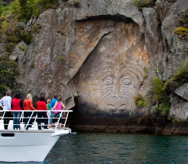 Group of people on boat looking at large Maori carving on rock face by water