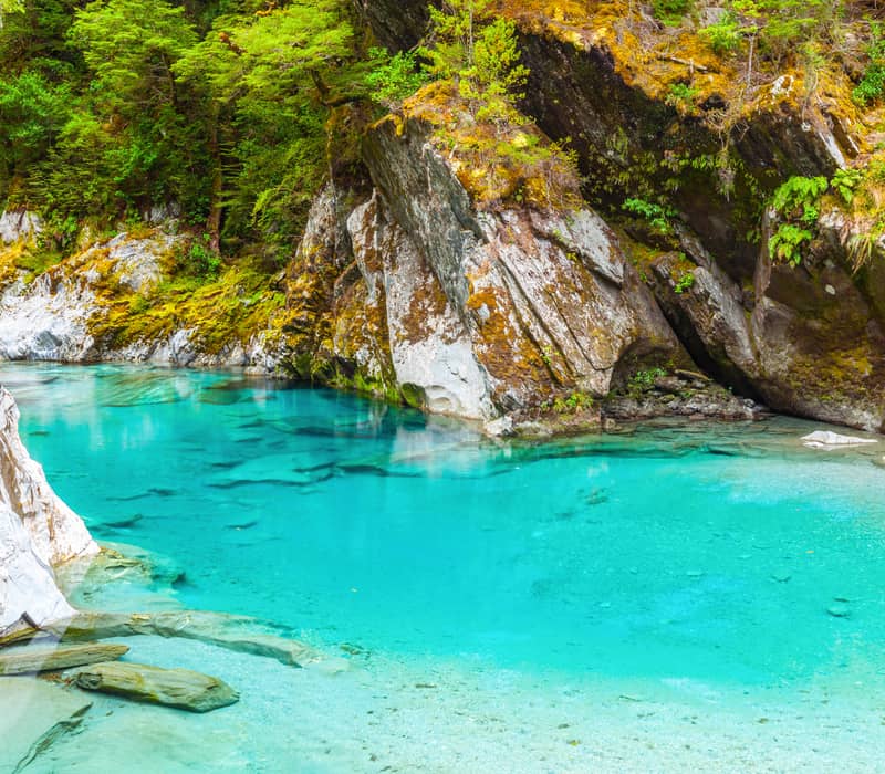Crystal clear blue water flowing through forested rocky river with moss-covered cliffs.