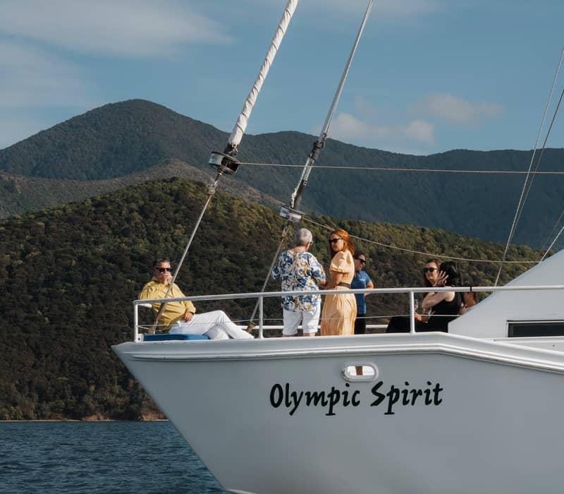People on a sailboat named Olympic Spirit with mountains and water in the background on a sunny day.