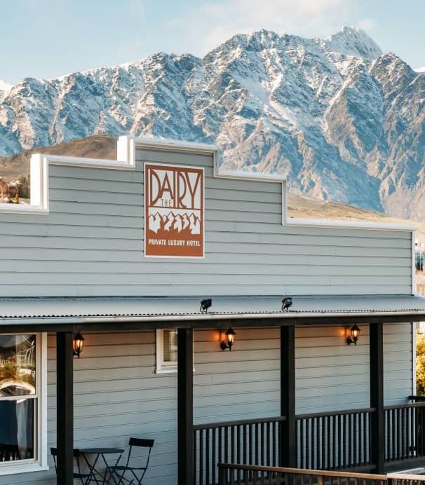 Hotel building with mountain backdrop and village nearby under clear blue sky.