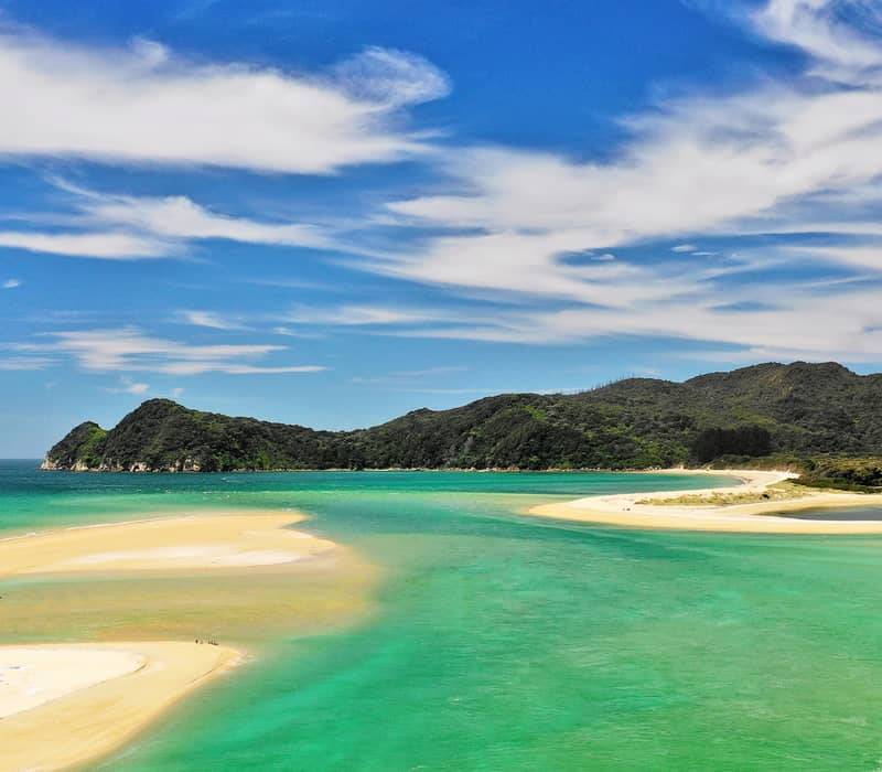 Sandy beach with turquoise water, green hills, and blue sky with clouds.
