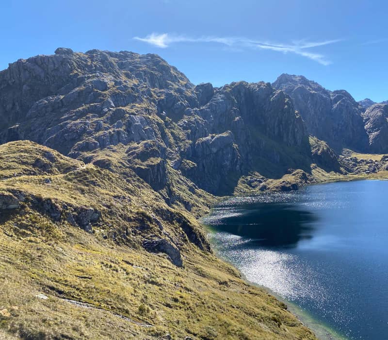 Mountain lake with rocky grassy slopes and hikers on a trail on a sunny day.