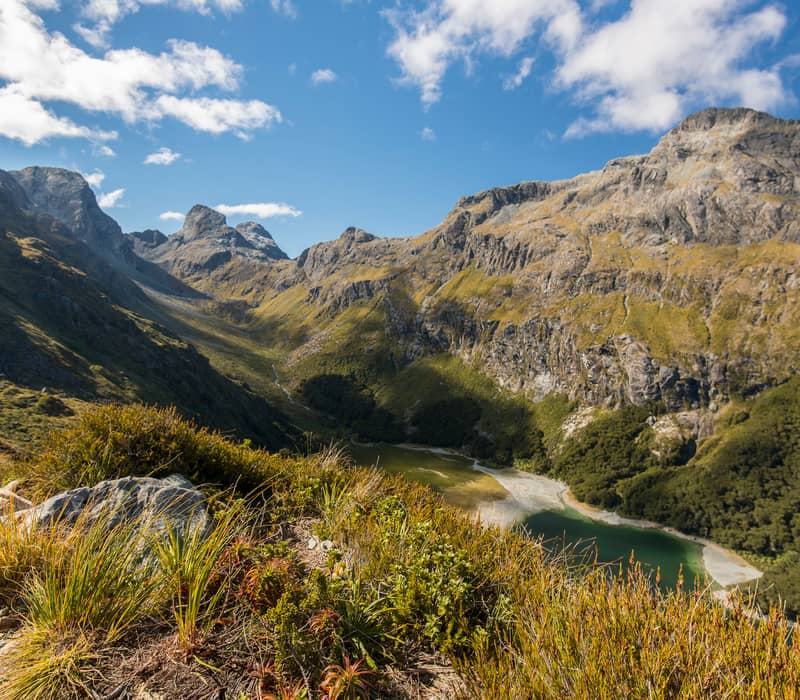 Mountain valley with a lake, green bushes, and blue sky with clouds.