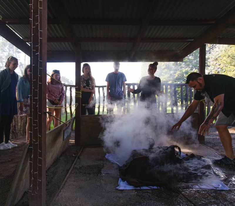A group of people watch steam rising from food being cooked in a hangi pit under a shelter.