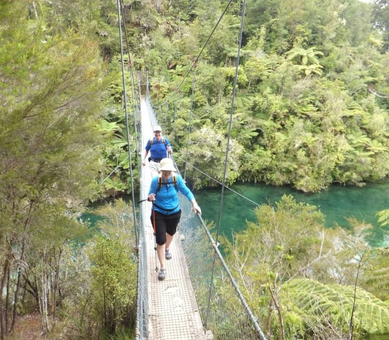 Two hikers walking on a suspension bridge over a green river amid dense forest.