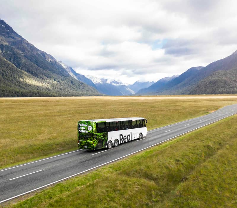 Tour bus driving on road through grassy valley with mountains and cloudy sky