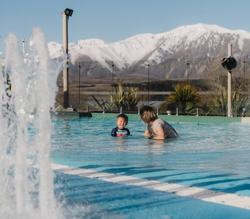 Adult and child swimming in outdoor pool with water fountain and snowy mountains behind.