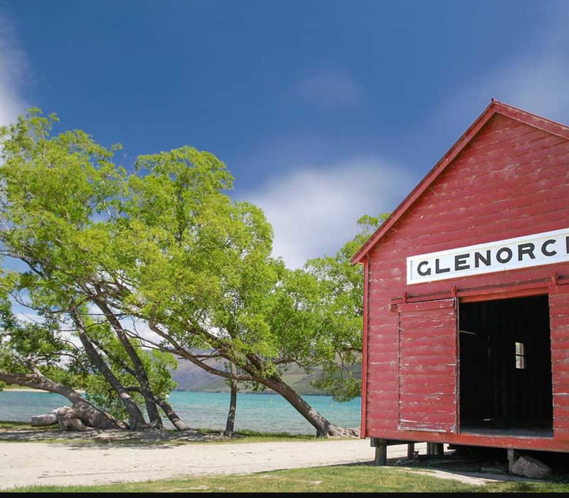 Red wooden shed labeled Glenorchy by a lake with trees and clear blue sky in the background.