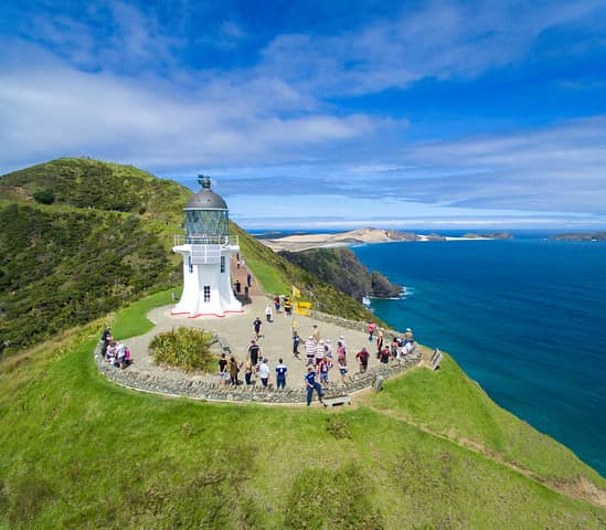 Lighthouse on a grassy hill overlooking the ocean with tourists around it on a clear day.