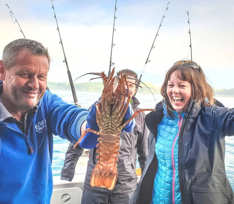 Three people on a boat holding up a large lobster with fishing rods and ocean in the background.