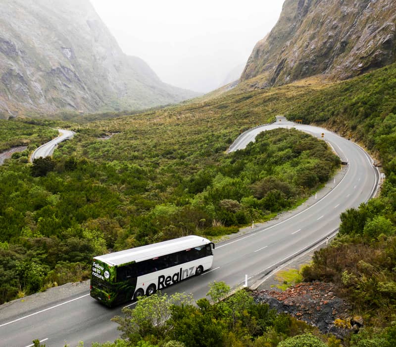 Tour bus on winding road through green valley with tall rocky mountains in the background.