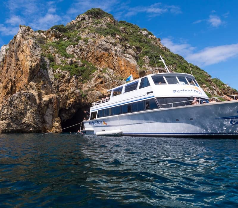 Tour boat anchored by a rocky island with cliffs in clear blue ocean water under a bright sky.