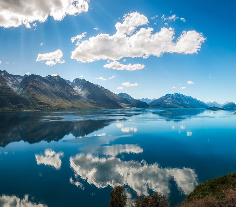 Clear lake reflecting rugged mountains and white clouds under a bright blue sky in Glenorchy.