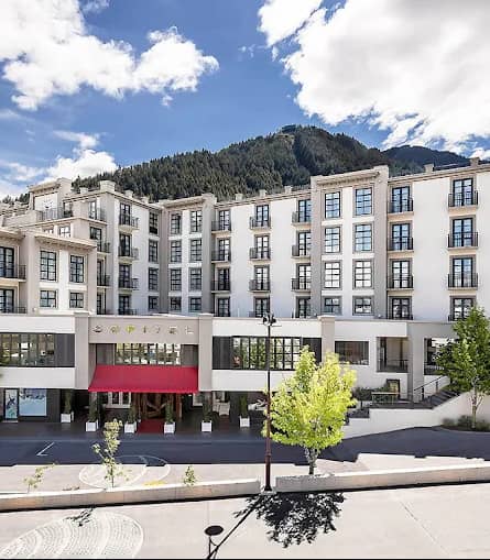 Modern white hotel building with mountain backdrop and red entrance canopy under a partly cloudy sky.