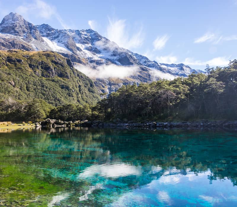 Clear lake with forest and snow-capped mountain reflections under blue sky