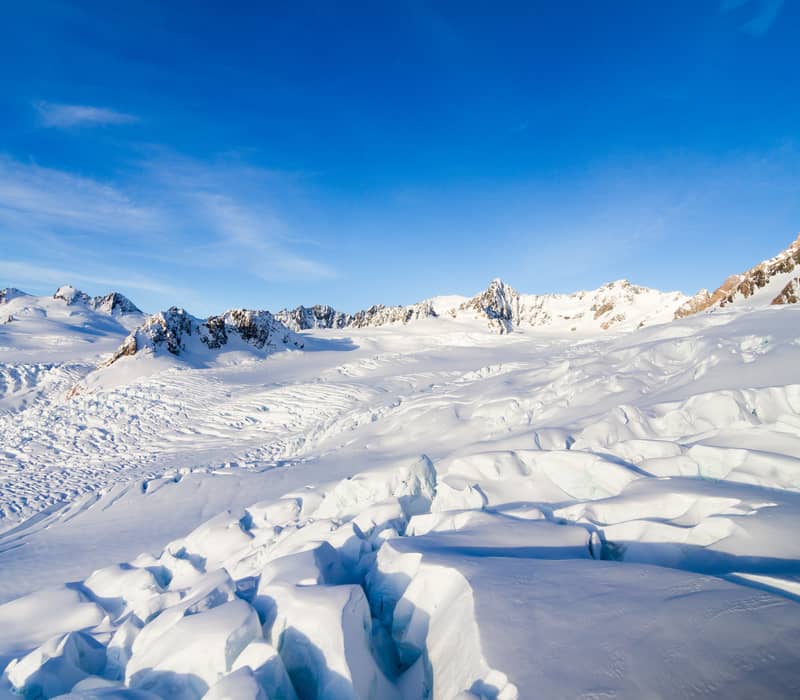 An expansive aerial view of the jagged ice falls and snow-covered névé of Fox Glacier under a bright blue sky.