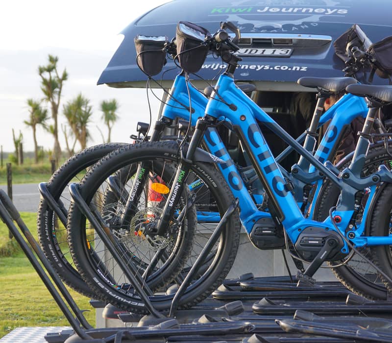 Blue electric bikes on a rack attached to a Kiwi Journeys vehicle outdoors.