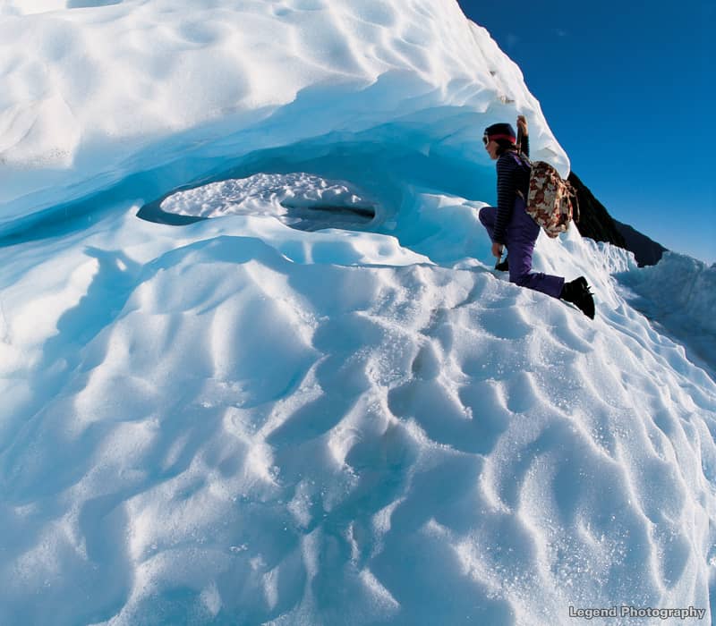 A hiker in purple gear and a backpack explores a glowing blue ice cave and dramatic ridges on the surface of Fox Glacier.