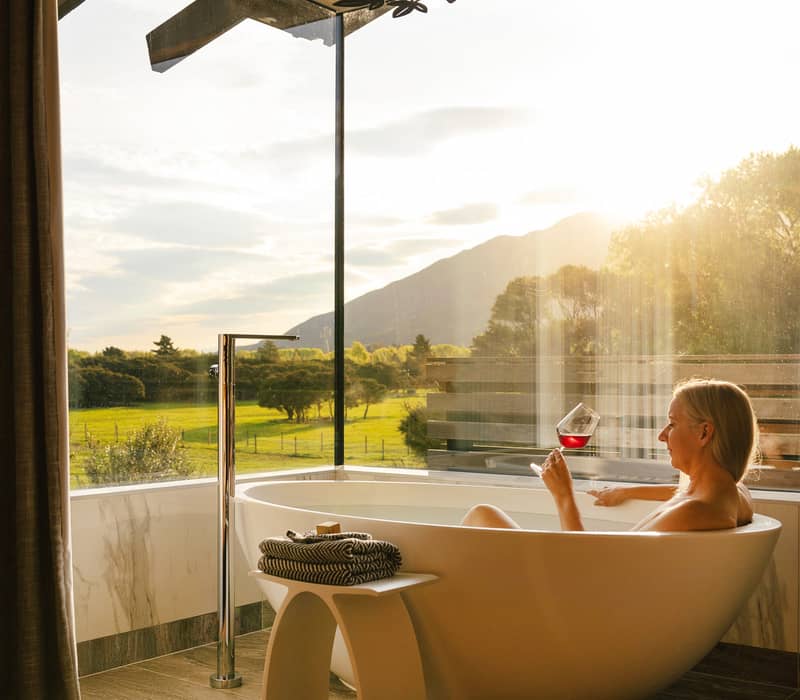 Woman in bathtub holding wine glass by window with mountain view and sunlight.
