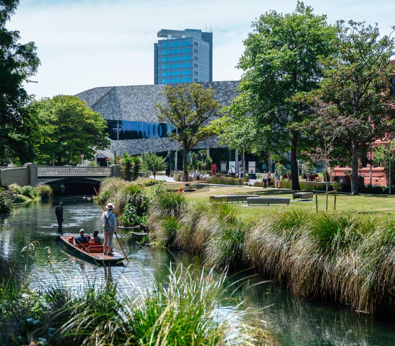People punting on a calm river with green trees, grass, and urban buildings in Christchurch.