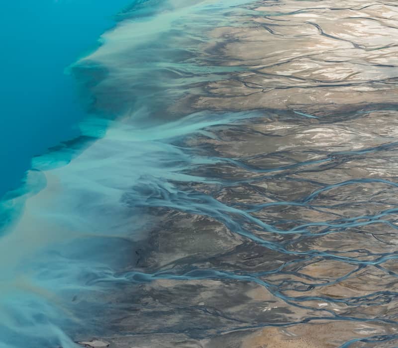 Aerial view of braided river channels flowing into bright blue water with sediment patterns.