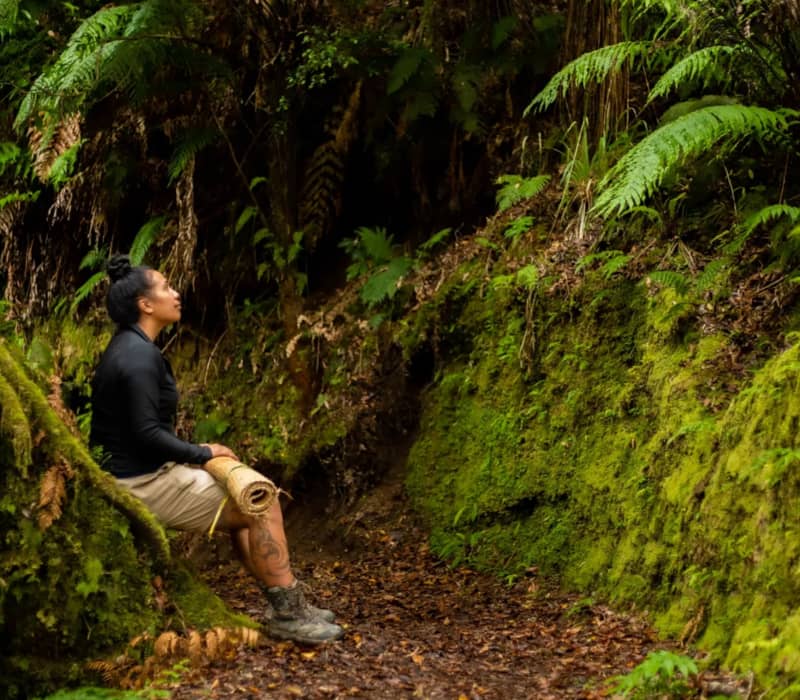 A Māori woman sits on a moss-covered tree root in a dense, green native forest, holding a woven flax mat (kete) and looking up toward the canopy.
