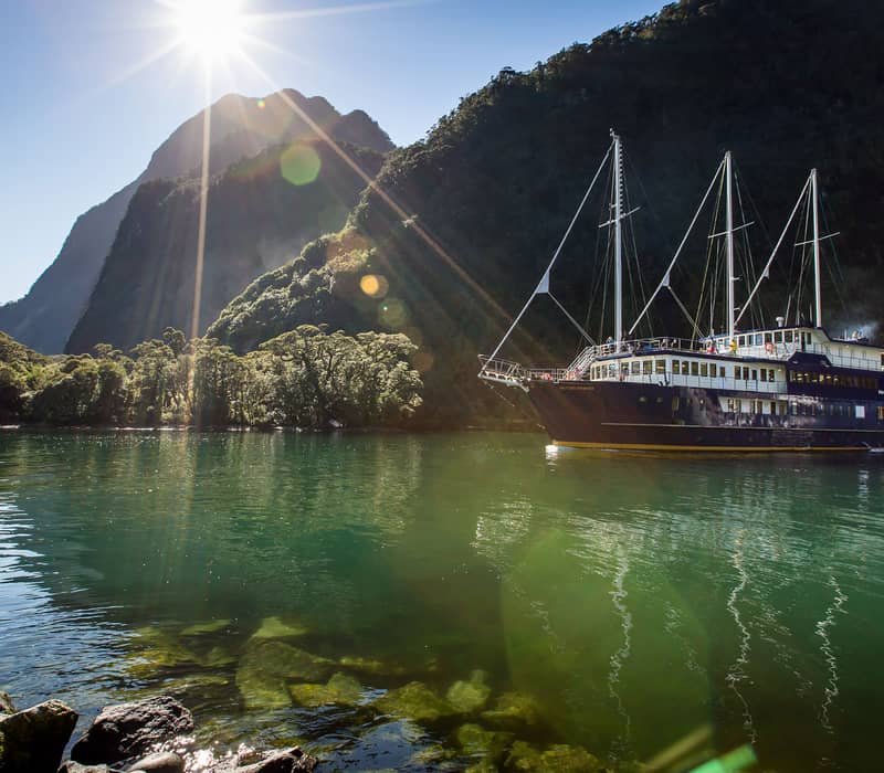 Sailboat on calm water with green mountains and bright sunlight in the background.