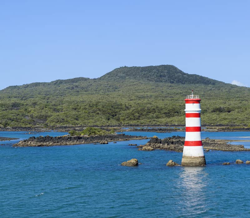 Red and white striped lighthouse in blue water near a green volcanic island with rocky shoreline.