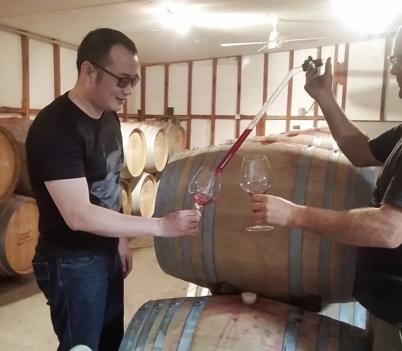 Two men tasting red wine from a barrel in a cellar filled with wooden barrels.