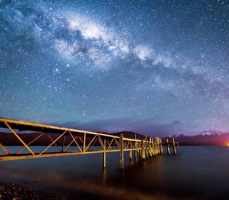 The Milky Way galaxy glows brilliantly above a long wooden jetty stretching into the calm, dark waters of Lake Te Anau.