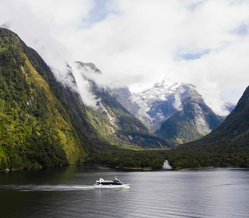 A white cruise boat sails on calm fjord water with towering green mountains and snow-capped peaks in the background.