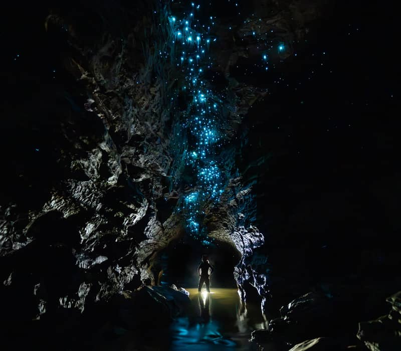 Silhouette of person standing inside a dark glowworm cave with blue glowing lights on the ceiling and reflections in the water.