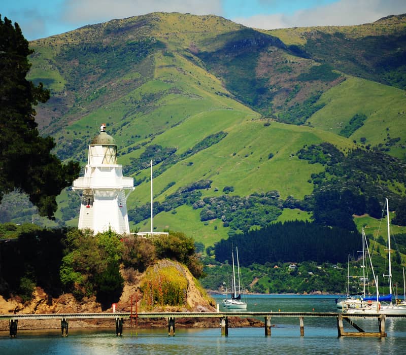 White lighthouse on a coastal hill with boats on the water and green hills in the background.