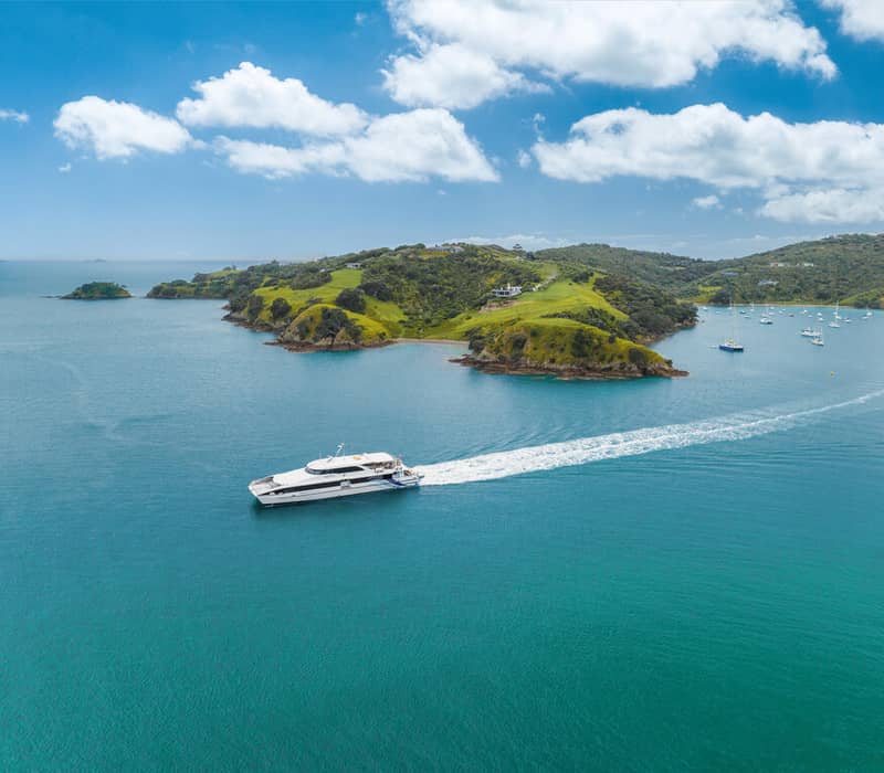 Ferry boat sailing near green hills of Waiheke Island with other boats anchored in calm blue water.