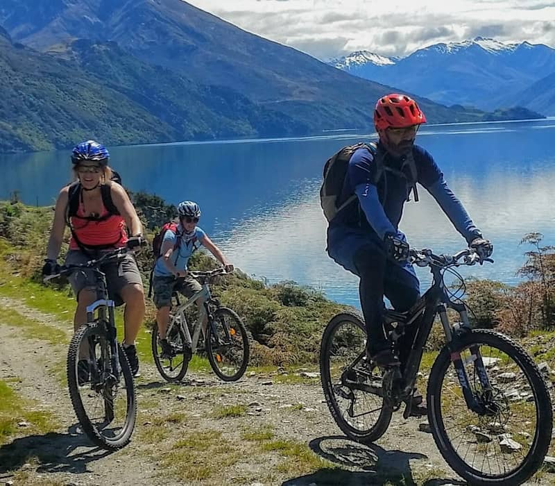Three cyclists mountain biking on a trail beside a lake with mountains behind them.