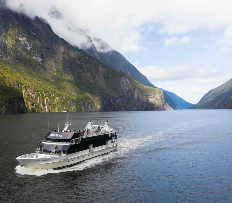 Cruise boat sailing through a mountain fjord with waterfalls and clouds above.