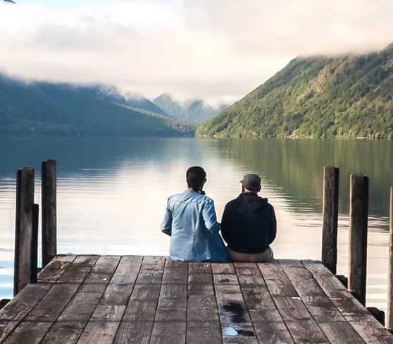 Two people sitting on a dock at Nelson Lake Rotoiti with misty hills.