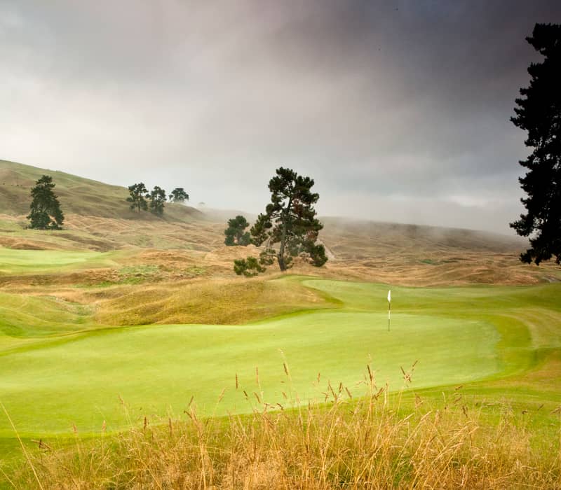 Green golf course with flag, trees and misty hills under overcast sky at Kinloch Golf 6th hole.