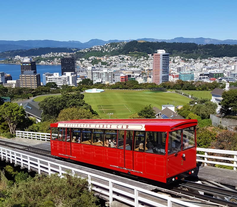 The bright red Wellington Cable Car travels down its tracks overlooking Wellington city, the harbor, and the surrounding hills under a clear blue sky.
