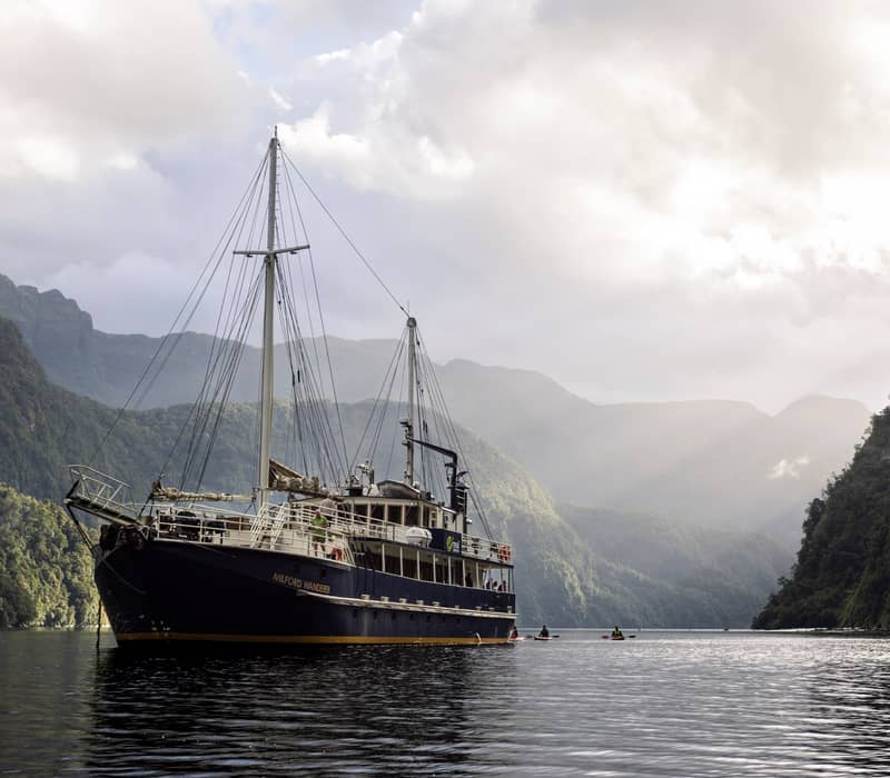 Boat on calm water between forest-covered mountains under cloudy sky with kayaks around