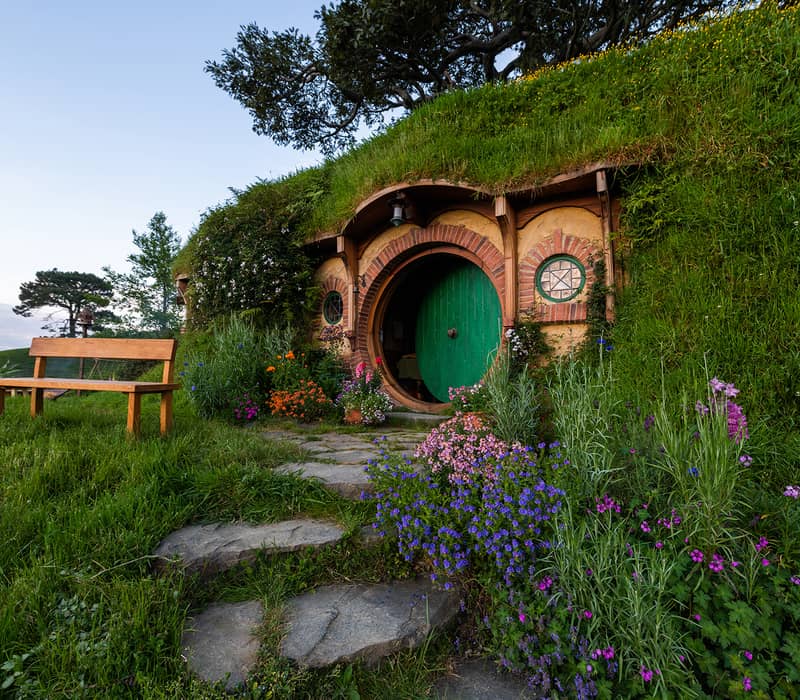 Hobbit hole with a green round door, surrounded by flowers and a wooden bench outside.