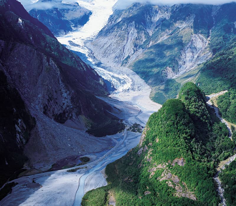 Aerial view of the Fox Glacier winding through a lush green valley with mountains and patches of snow.
