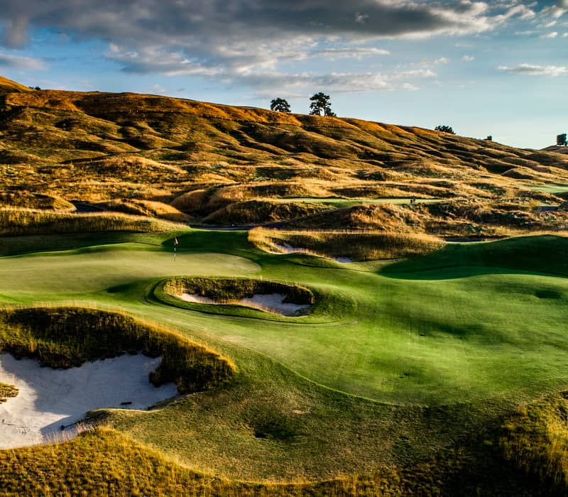 Golf course fairway with green, sand bunkers, and hills under cloudy sunset sky.