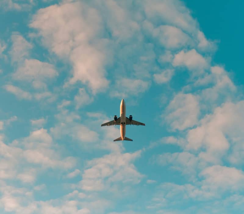 An airplane seen from directly below flying high in a bright blue sky scattered with fluffy white clouds.