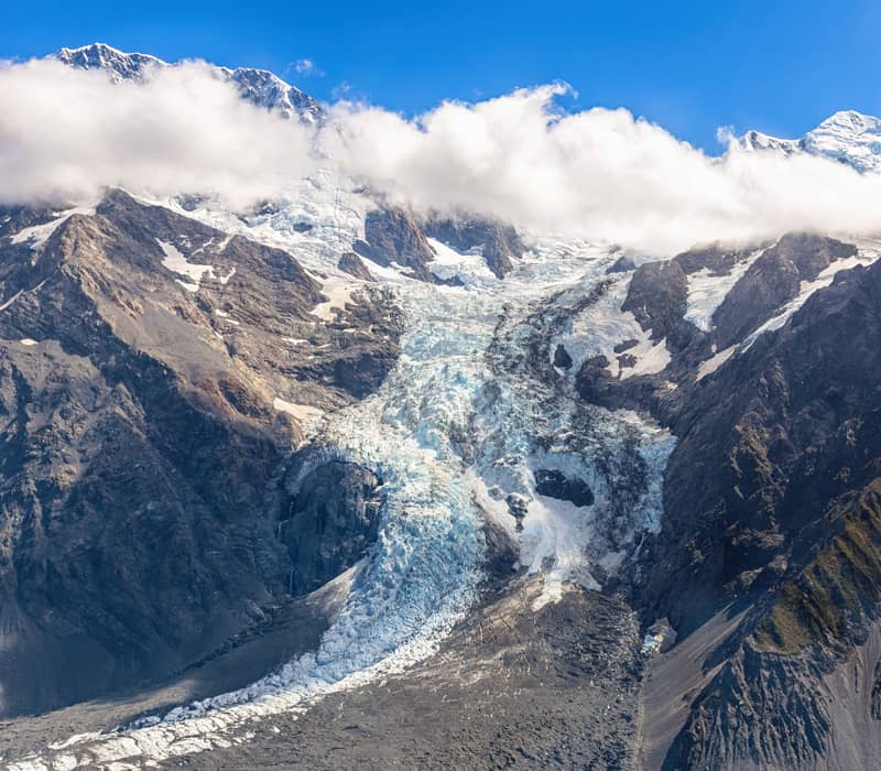 Snow-capped Fox Glacier and rocky mountains under blue sky with white clouds.