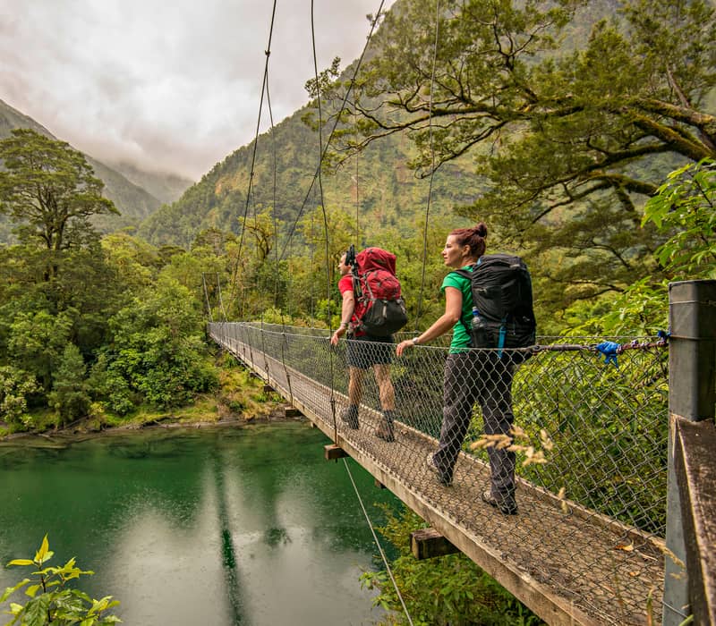 Two hikers with backpacks crossing a suspension bridge over a river in a forested mountain area.