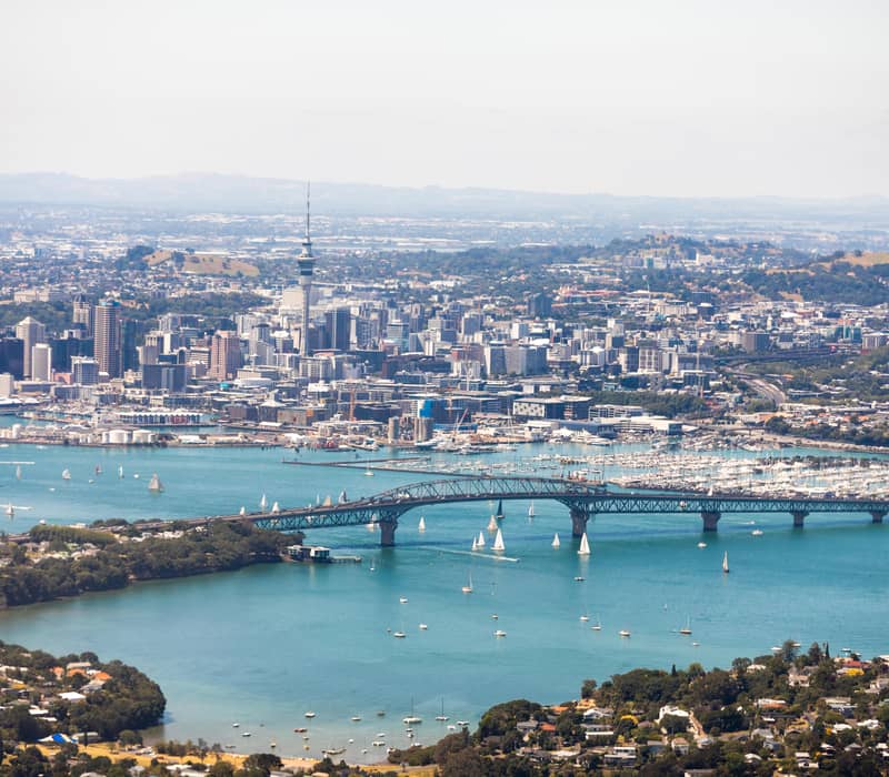 Auckland Harbour Bridge with city skyline and sailboats on blue water under clear sky.