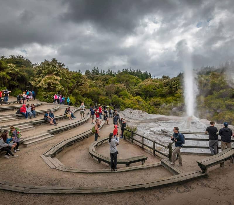 Crowd of tourists watching an erupting geyser at Waiotapu Thermal Wonderland from wooden benches.