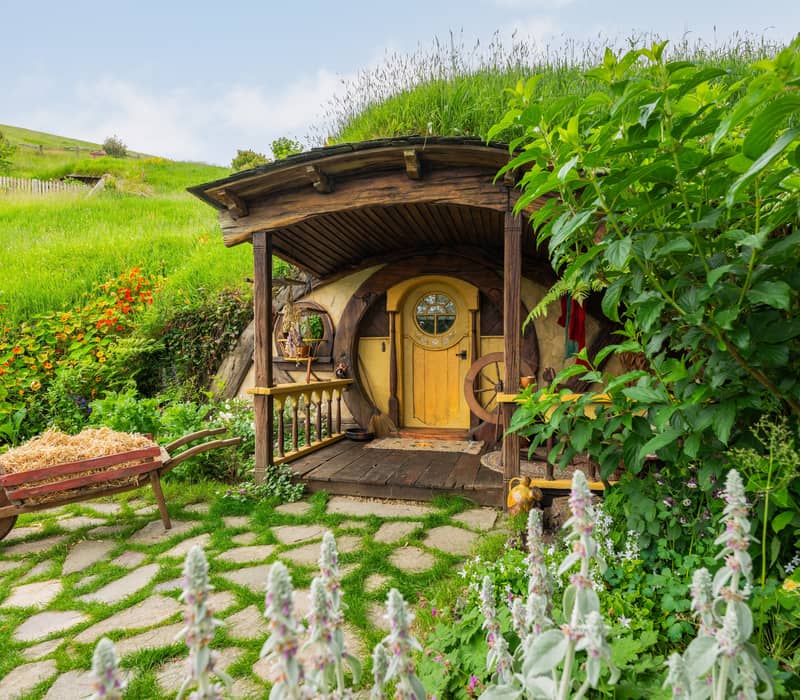 Hobbit house with round yellow door and wooden porch surrounded by greenery and flowers with a wheelbarrow in front.