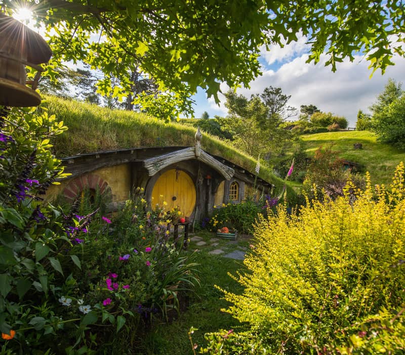 Hobbit hole with yellow door surrounded by vibrant garden and greenery under sunny sky.